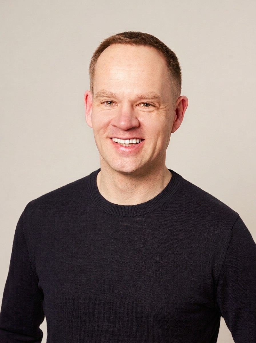 Portrait von Axel M. Heinz: lächelnd, in dunklem Pullover vor hellem Hintergrund