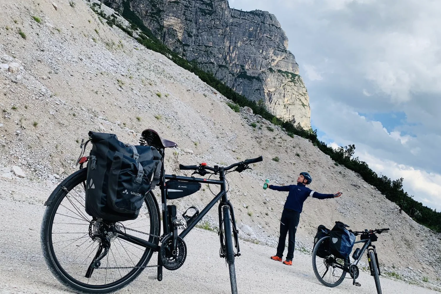Zwei bepackte Fahrräder auf einem Schotterweg unter einer steilen Felswand, daneben ein Radfahrer mit ausgebreiteten Armen