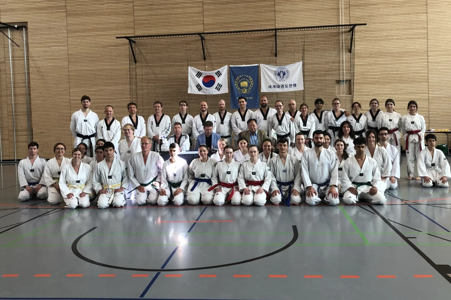 Gruppenfoto nach einer Taekwondo-Prüfung: rund 50 Sportlerinnen und Sportler in weißen Anzügen in einer Sporthalle